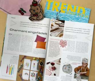 zu sehen ist die aufgeschlagene Ausgabe des Fachmagazins Trend and Style, in dem man den Artikel sieht sowie 2 bestickte Produkte